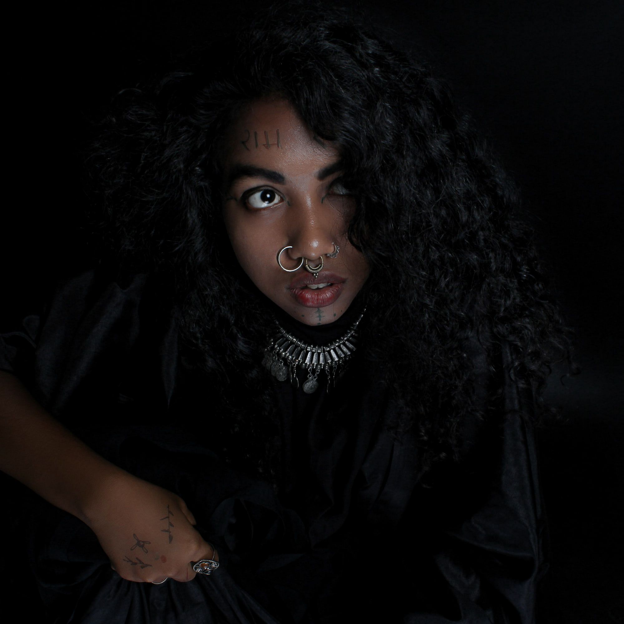 A woman in a black dress sits in a dark studio, her lips a deep garnet red.